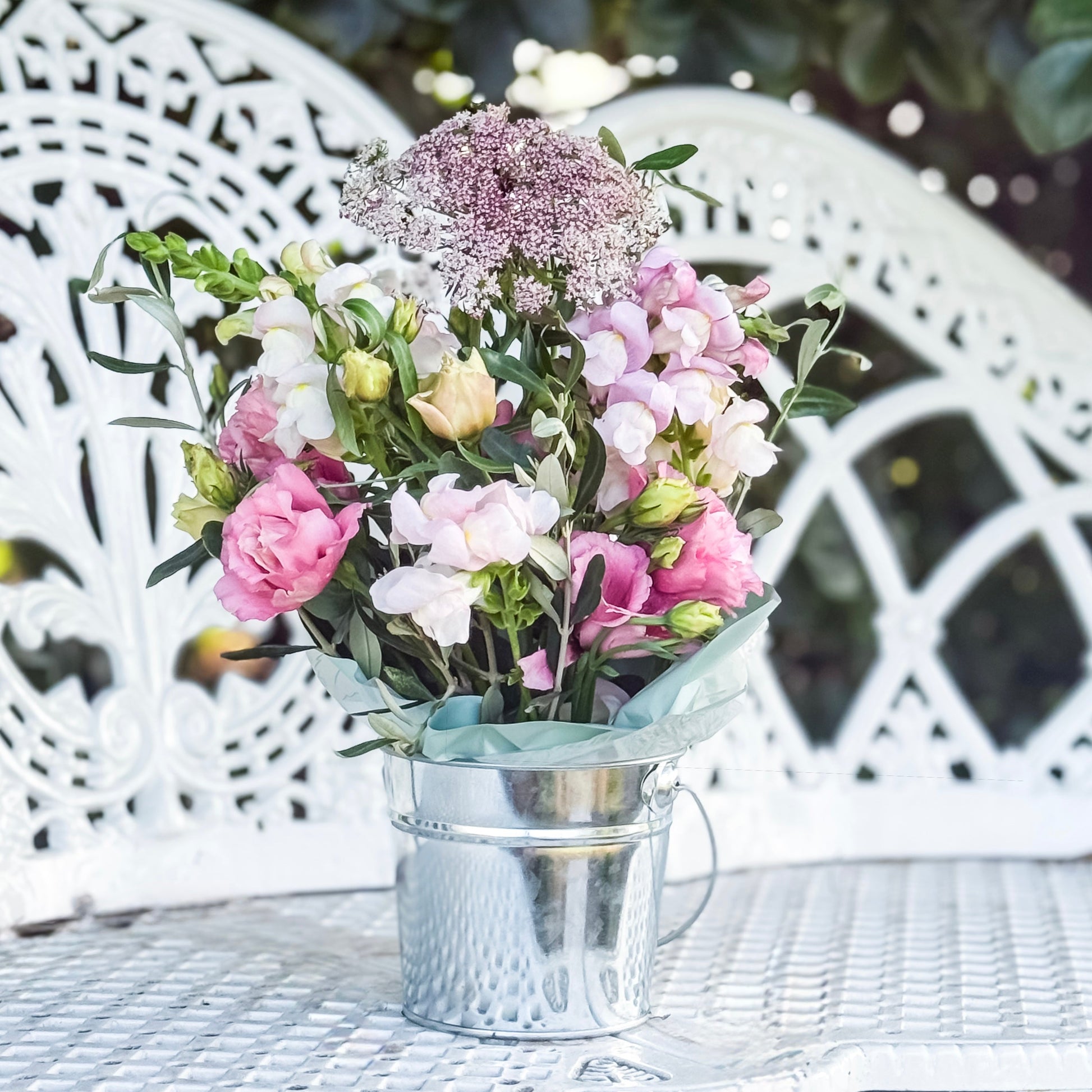 Bouquet of flowers in a metal container on a white wicker table outdoors.