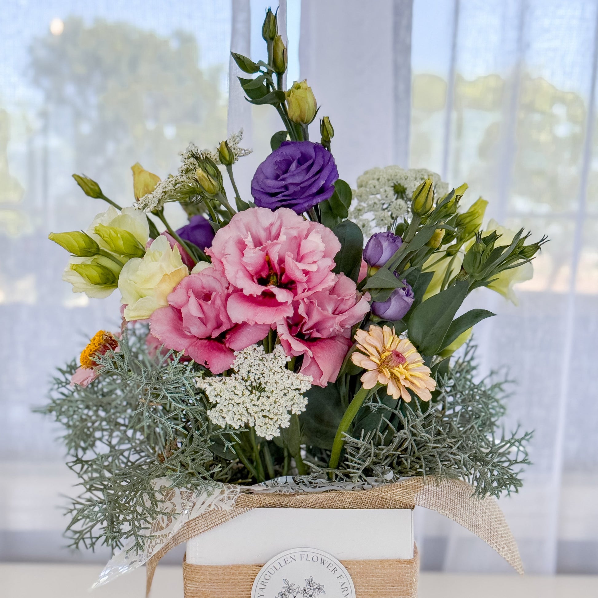 Colourful flower arrangement in a box with a visible brand label, set against a blurred outdoor background.