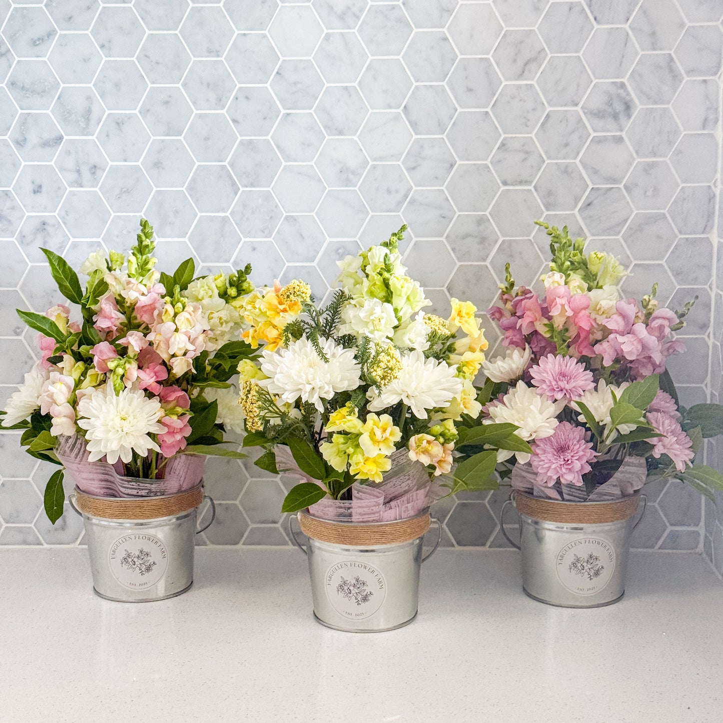Three flower arrangements in metal buckets against a hexagonal tile wall.